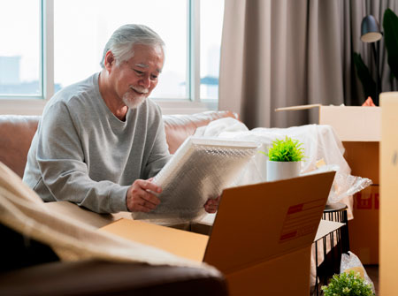 man sitting in a room with moving boxes smiling at a wrapped picture frame in his hands