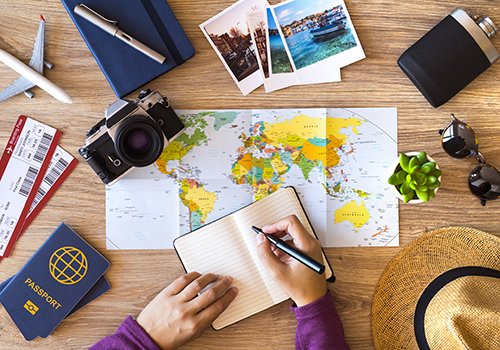 Woman making a list for her travel, on a wooden desktop with travel accessories.