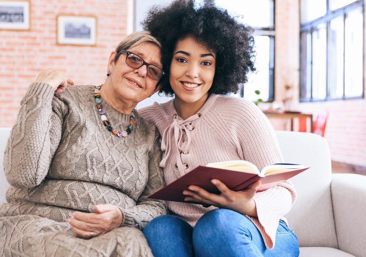 young woman holding an open book whiling sitting on the couch with her senior mother