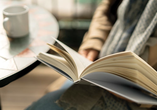 close up of a person sitting at their breakfast table with a cup of coffee, paging through a hardcover book