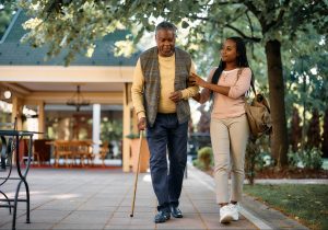 senior man with walking cane and his daughter taking a walk through the park of a senior living home.