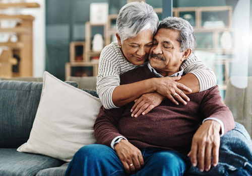 senior man sitting on a loveseat with his wife smiling and embracing him