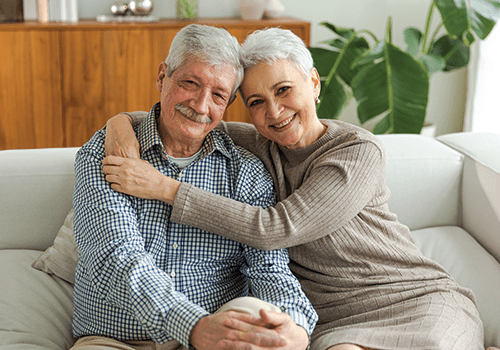 senior woman with her arms around her husband as they sit in their living room